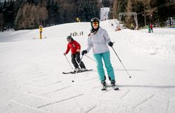 Zwei Skifahrer in Winterkleidung fahren auf einer präparierten Skipiste mit Wald im Hintergrund