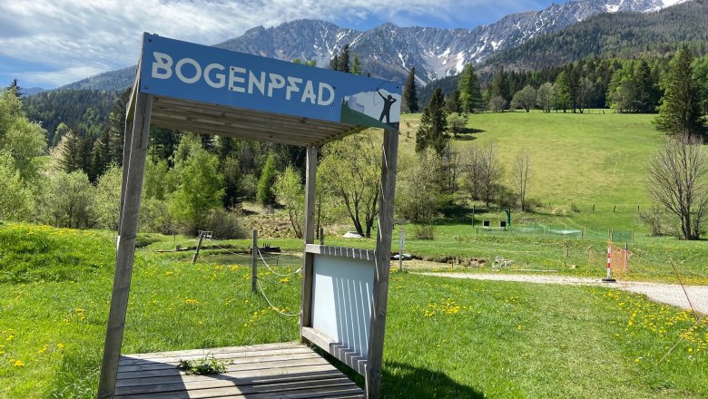 Holzplattform mit Schild 'Bogenpfad' vor einer Berglandschaft.