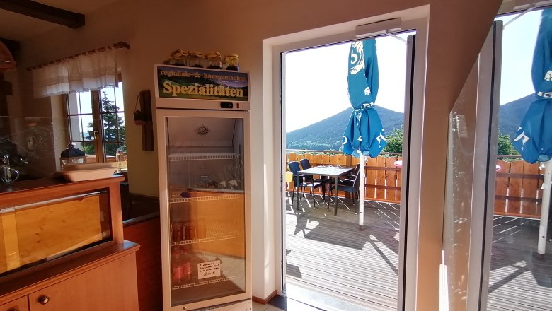 Blick von innen auf eine Terrasse mit Sonnenschirmen und Bergen im Hintergrund.