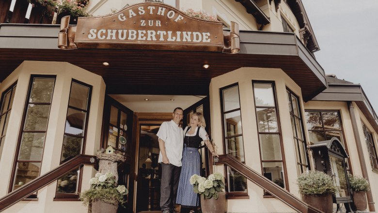 Eingang des Gasthofs Zur Schubertlinde mit zwei Personen in traditioneller Kleidung.