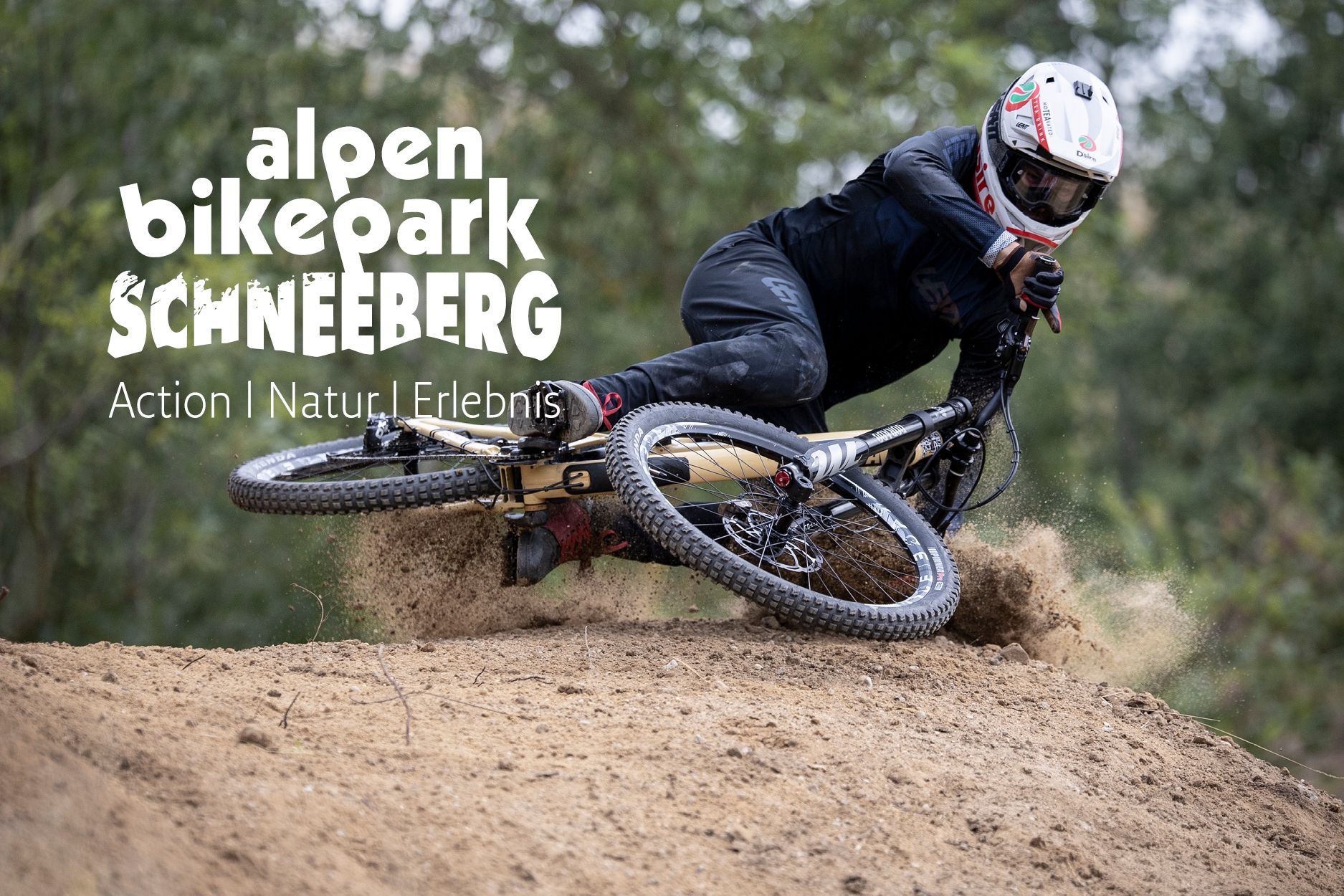 Eine Person mit Helm fährt auf einem Mountainbike durch eine sandige Kurve. Aufgewirbelter Sand ist sichtbar, im Hintergrund stehen Bäume.