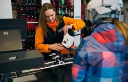 Verkäuferin mit orangefarbenem Pullover montiert Skischuh an Skibindung vor Kundin in Skihelm und bunter Jacke