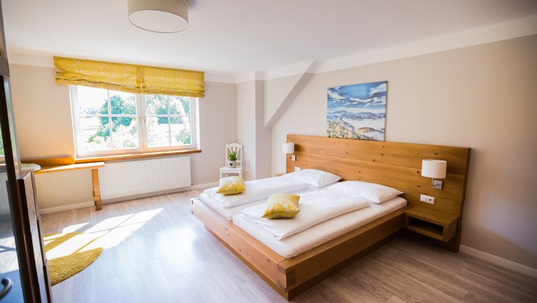 Helles Doppelzimmer mit Holzbett, gelben Kissen und gro&szlig;em Fenster.