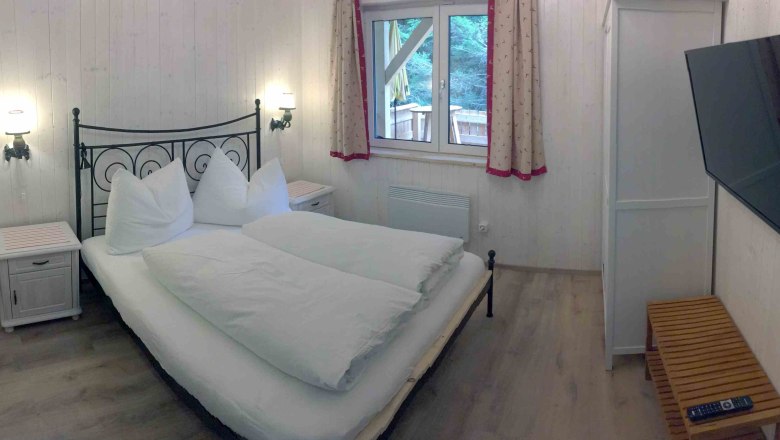 Schlafzimmer in einem Ferienhaus mit Doppelbett, TV und Fenster mit Vorh&auml;ngen.