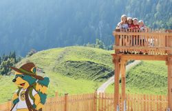 Vier Kinder stehen auf einem hölzernen Aussichtsturm vor einer grünen Berglandschaft mit Wald und Nebel.