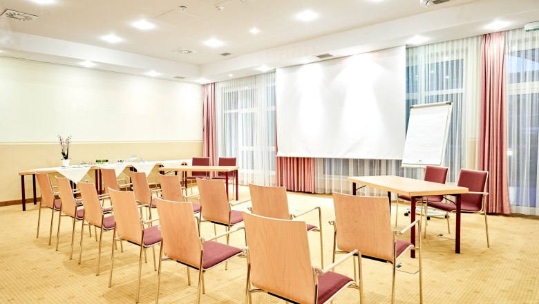 Ein heller Seminarraum mit St&uuml;hlen, Tischen, einem Flipchart und einem Projektionsschirm.