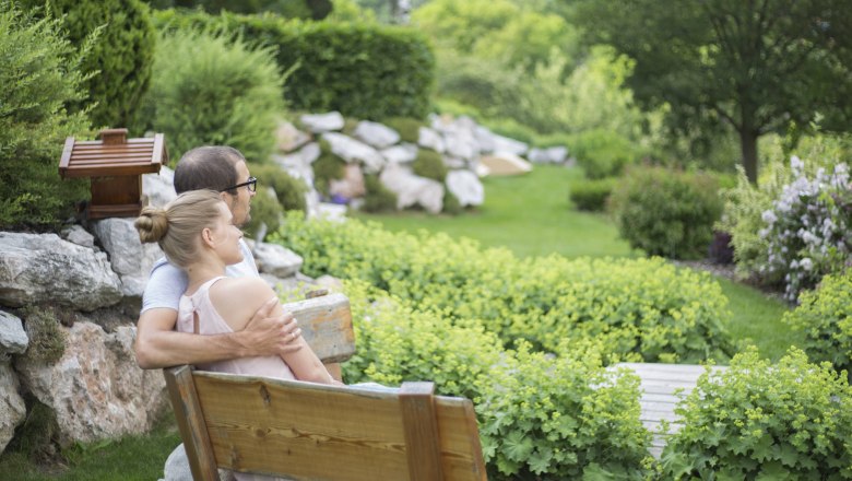 Ein Paar sitzt auf einer Holzbank in einem gr&uuml;nen Garten mit B&uuml;schen und B&auml;umen.