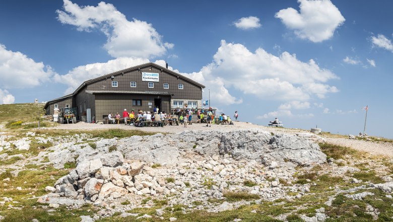 Fischerh&uuml;tte auf dem Schneeberg mit Wanderern und blauem Himmel.