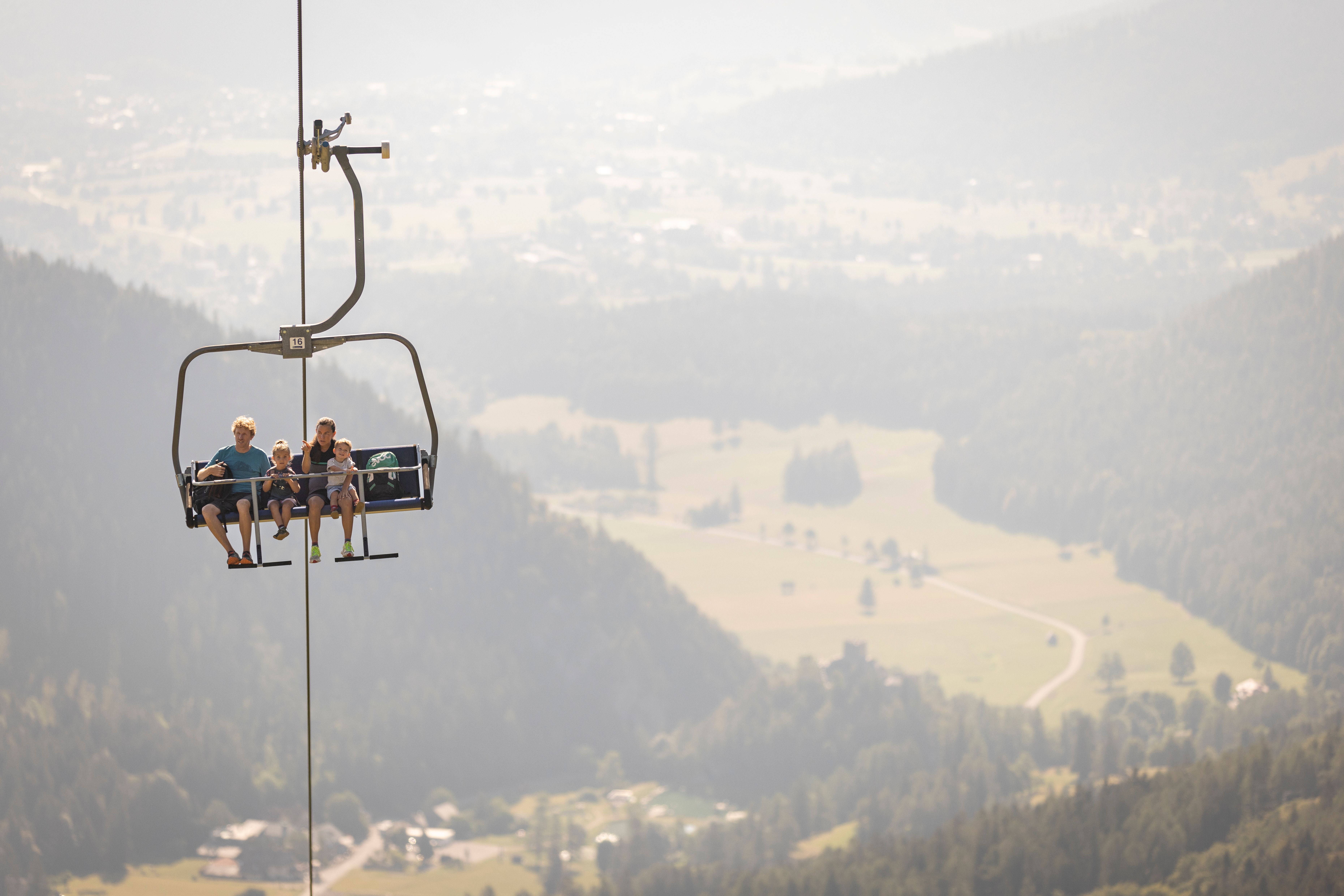 Vier Personen sitzen auf einem Sessellift über bewaldeten Hügeln mit weiter Landschaft im Hintergrund