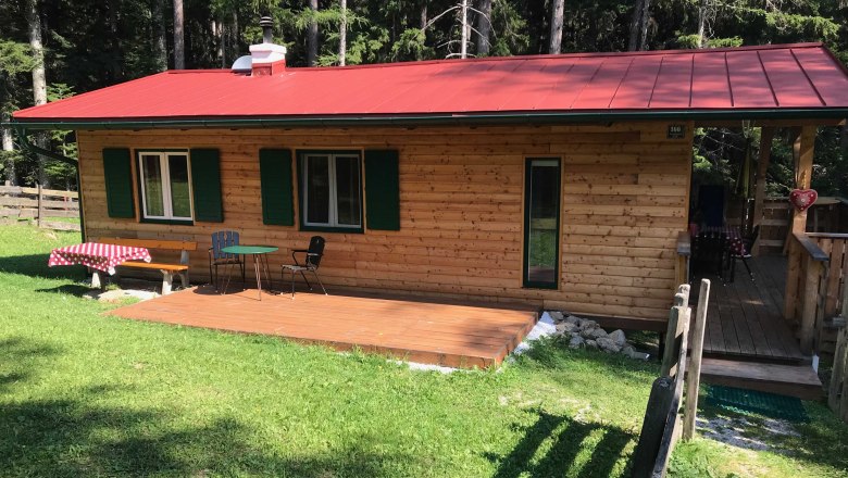 Ein kleines Holzferienhaus mit rotem Dach, Terrasse und Gartenm&ouml;beln, umgeben von Wald.
