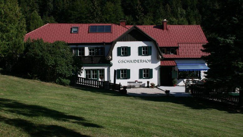 Ein traditionelles Geb&auml;ude mit rotem Dach und gr&uuml;nen Fensterl&auml;den, umgeben von B&auml;umen und Wiese, mit dem Schriftzug 'Gschaiderhof'.