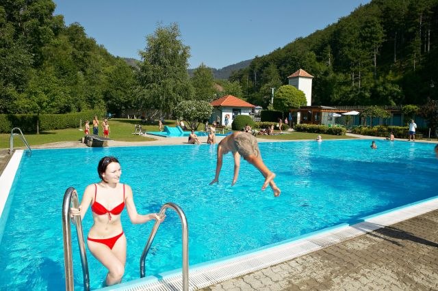 An outdoor pool with people swimming and relaxing. A woman gets out of the water while a man jumps into the pool. Surrounded by trees and hills.