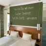 Ein Doppelzimmer mit zwei Betten, gr&uuml;ner Wand mit Spruch, Vorh&auml;ngen und einer Dusche mit Glaswand.