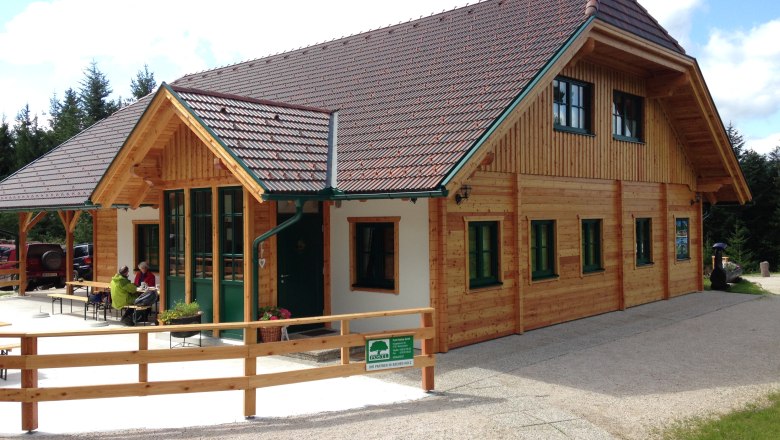 Eine Holzhütte mit grünem Dach und Fenstern, umgeben von Bäumen. Zwei Personen sitzen draußen an einem Tisch.