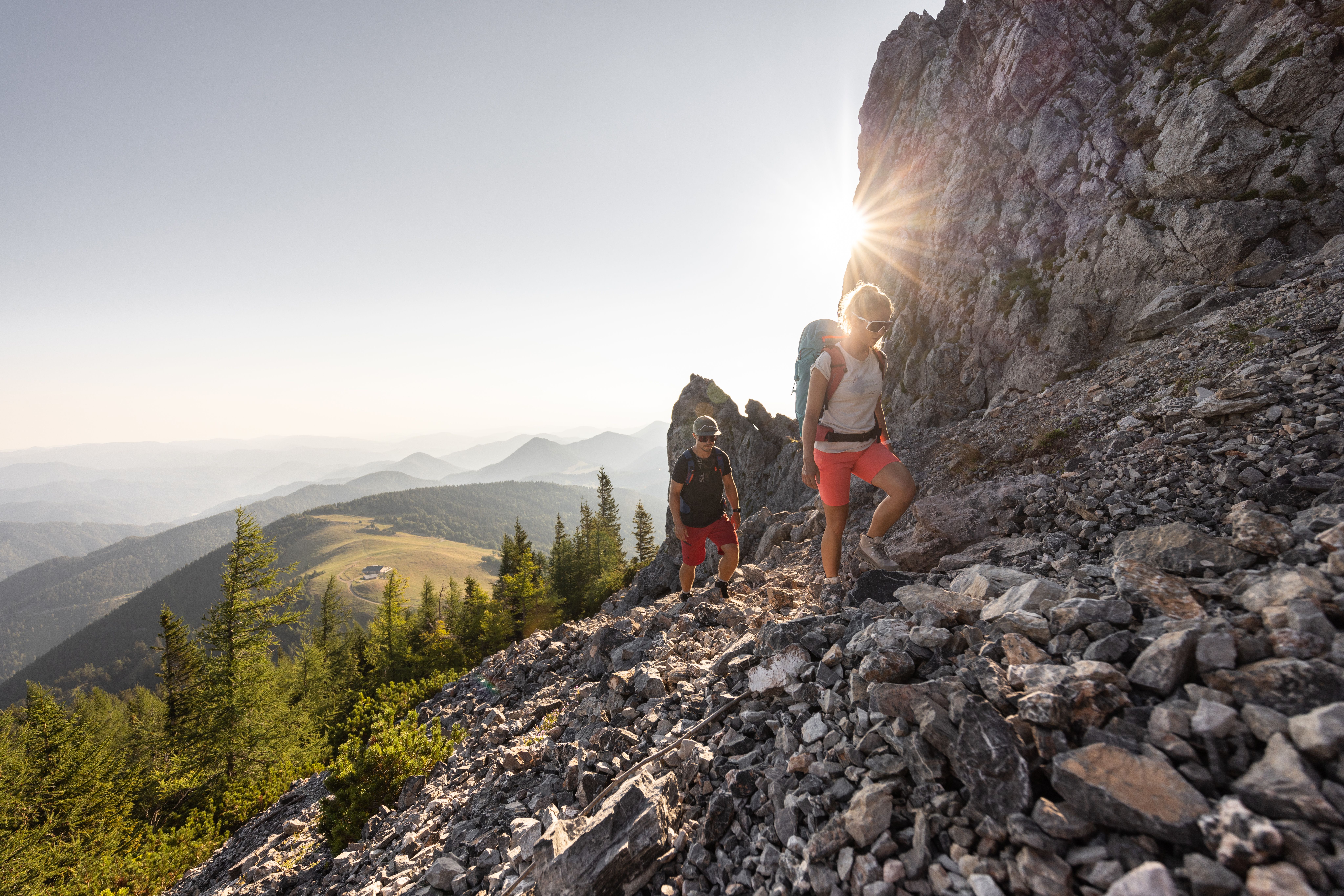 Zwei Wanderer mit Rucksäcken steigen auf steinigem Gebirgspfad bei Sonnenschein und Bergpanorama auf