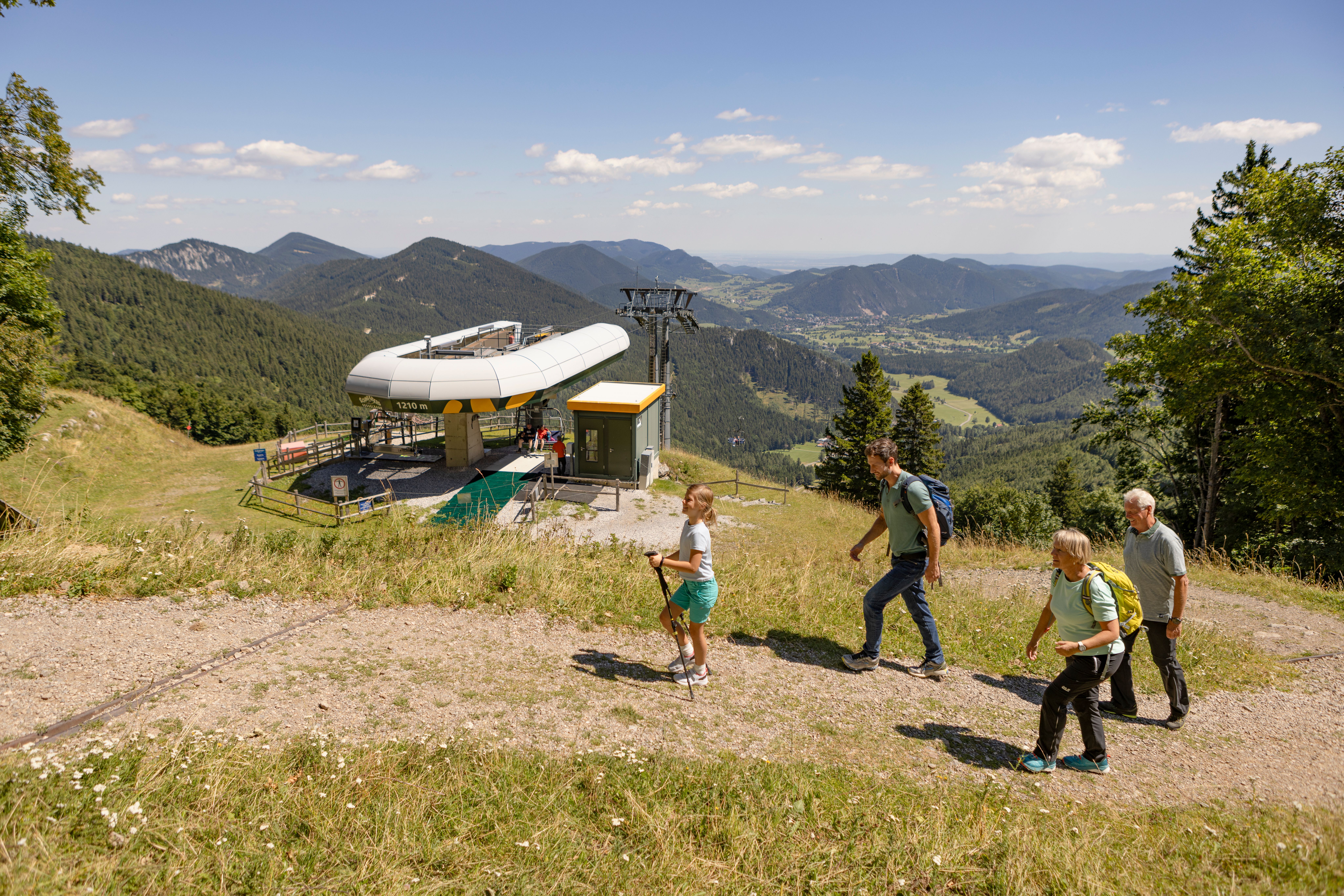Vier Wanderer mit Rucksäcken steigen auf einem Bergweg an einer Bergstation einer Seilbahn vorbei mit Blick auf bewaldete Berge und ein Tal unter blauem Himmel