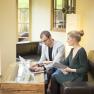 Two people work on a laptop in a cozy room with large windows.