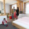 Familie in einem gem&uuml;tlichen Hotelzimmer mit Holzm&ouml;beln und rosa Bettw&auml;sche.