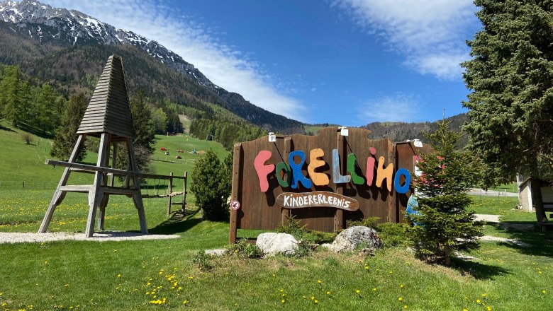 Eingang zum Forellino Kinderspielplatz mit Berglandschaft im Hintergrund.