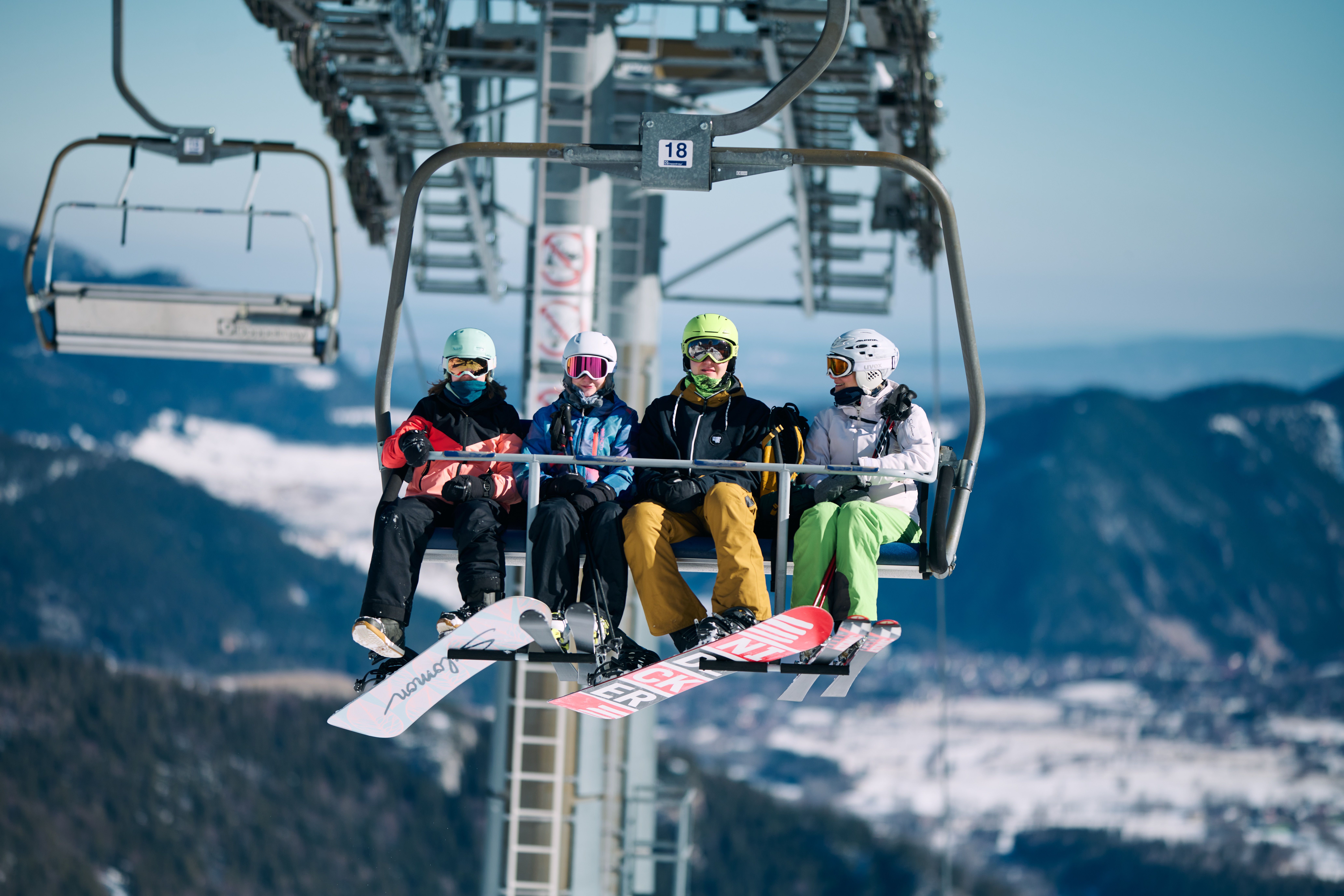 Vier Personen in Skibekleidung sitzen auf einem Skilift mit Skiern und Snowboards, im Hintergrund verschneite Berge und blauer Himmel