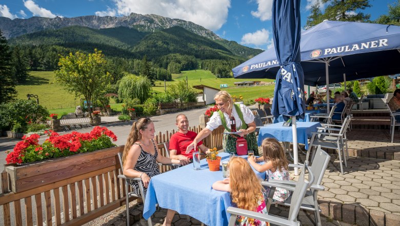 Familie auf sonniger Terrasse mit Bergen im Hintergrund, bedient von Kellnerin in traditioneller Kleidung.