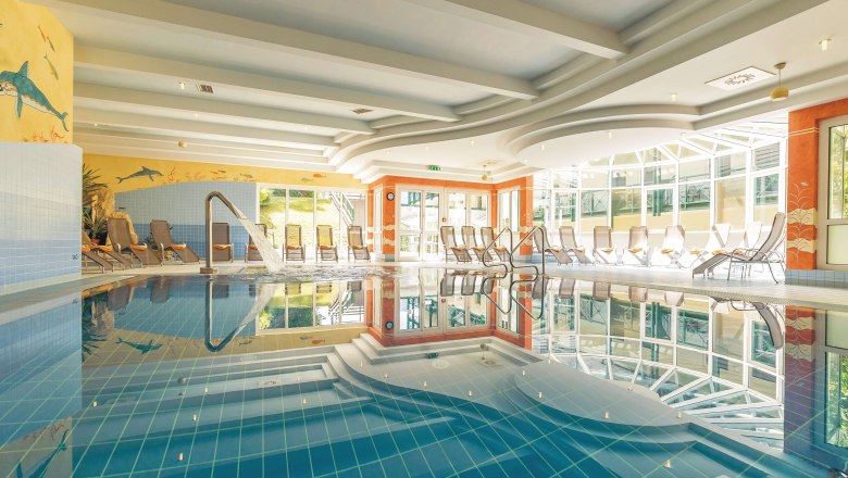 Ein helles Hallenbad mit Liegest&uuml;hlen und einem Wasserstrahl im Pool.