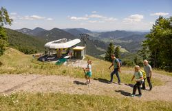Vier Wanderer mit Rucksäcken steigen auf einem Bergweg an einer Bergstation einer Seilbahn vorbei mit Blick auf bewaldete Berge und ein Tal unter blauem Himmel